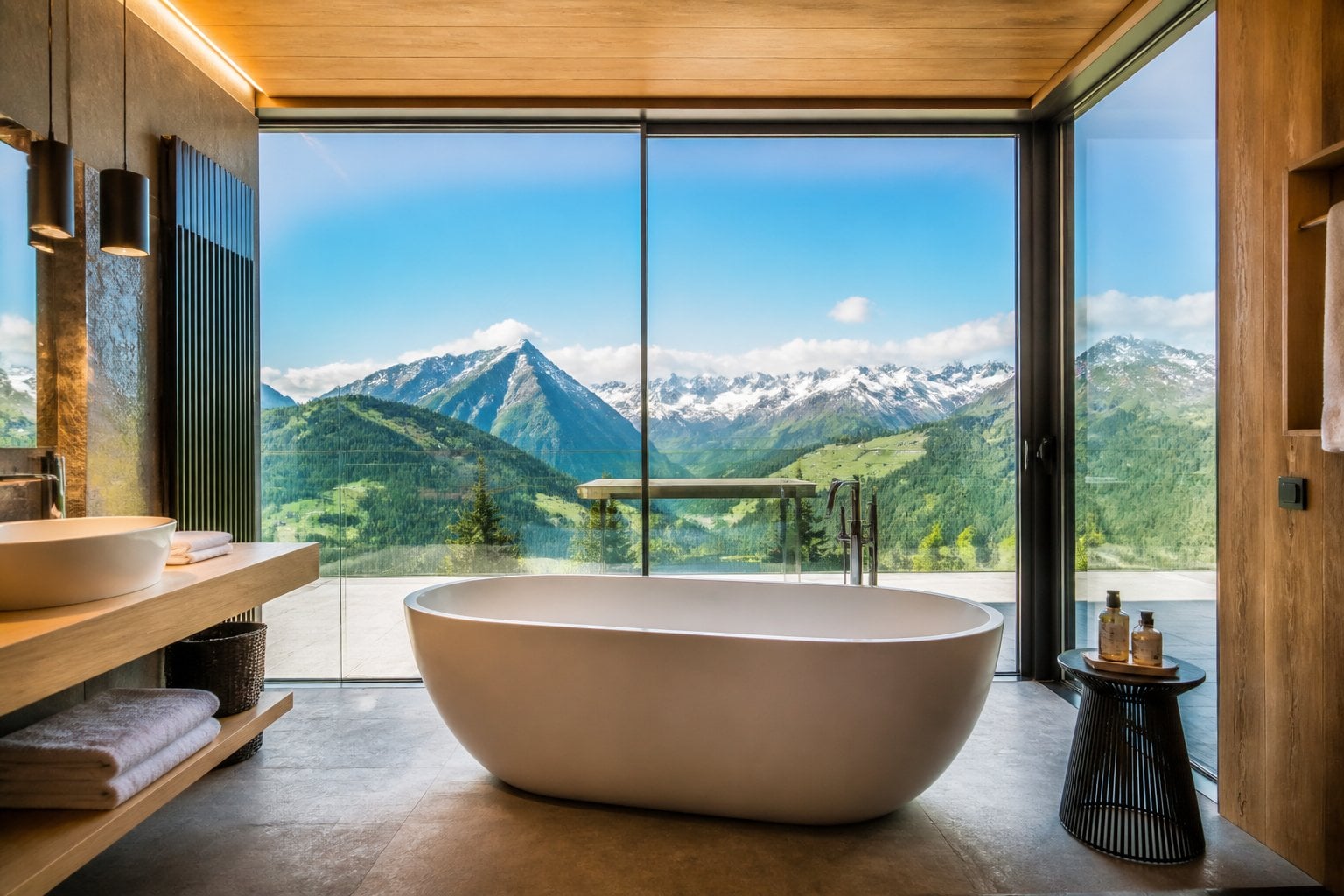 A modern bathroom with a window bathtub overlooking snow-capped mountains and green alpine scenery through large glass windows.