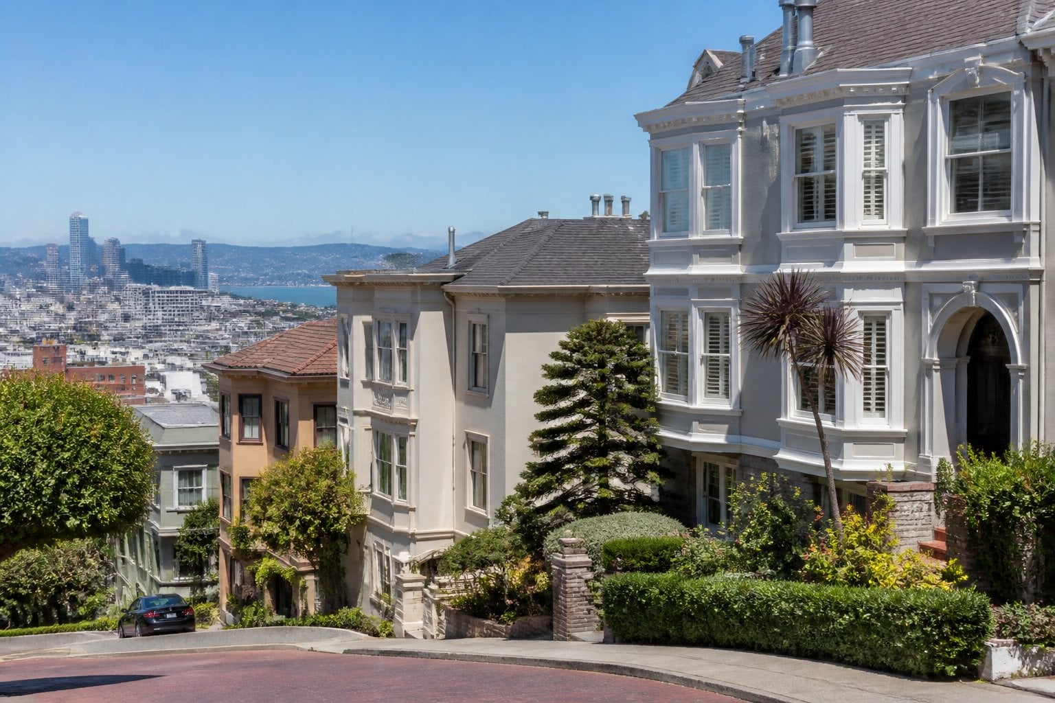 Upscale Victorian homes line a landscaped hillside street in San Francisco with the bay and distant hills in the background.