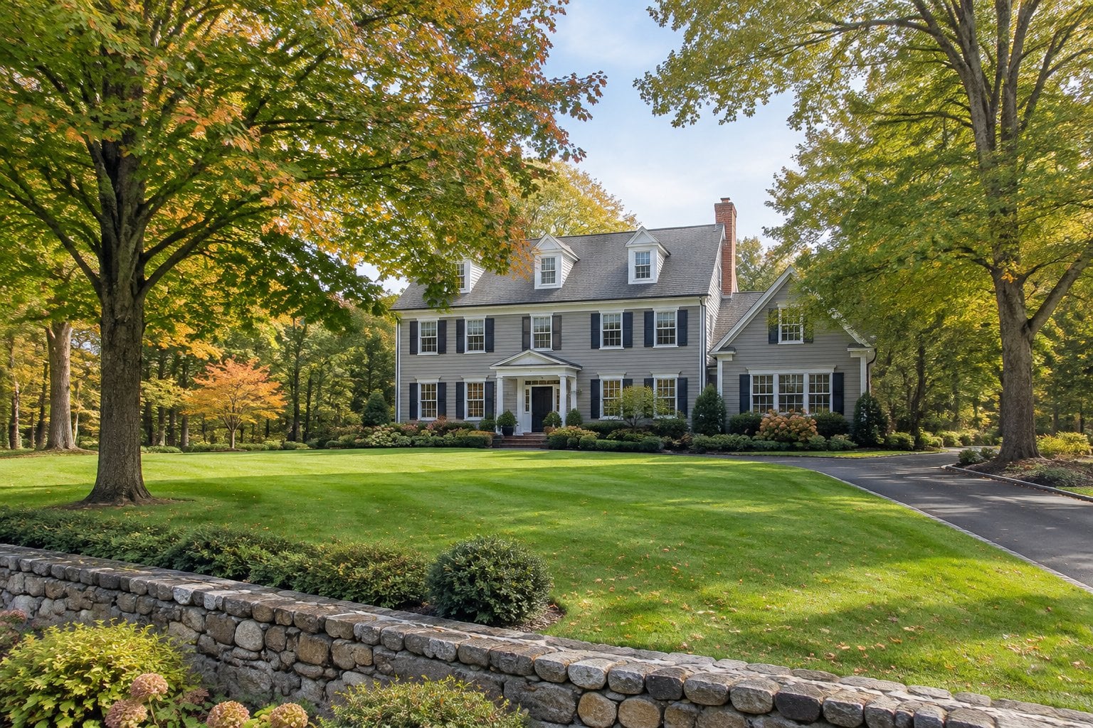 A large colonial-style home sits among mature trees and landscaped lawns in a quiet suburban neighborhood.